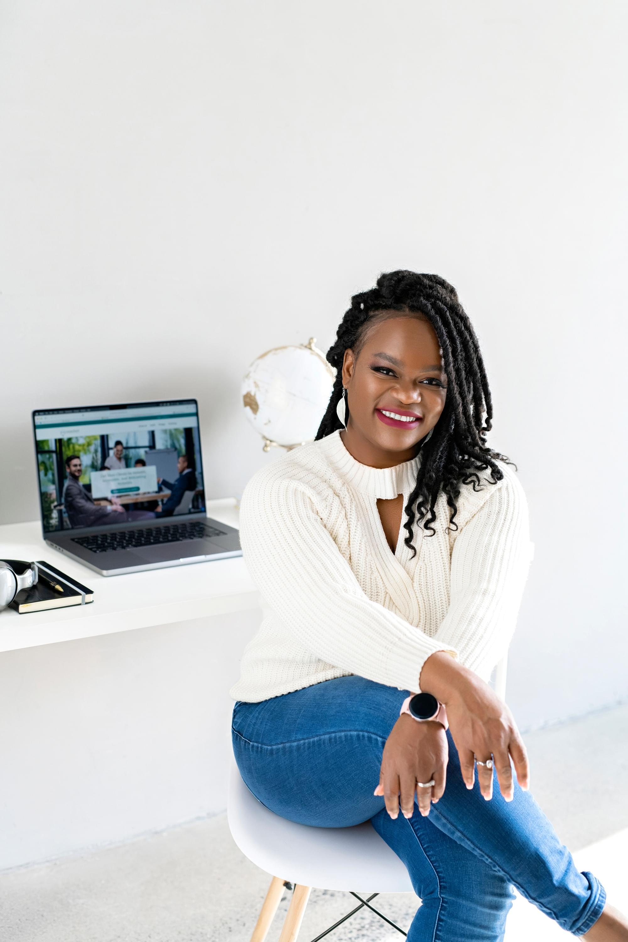 Flora Bazie, CEO of LInoraTech Inclusion, sits at a desk wearing a white sweater and smiles at camera. 